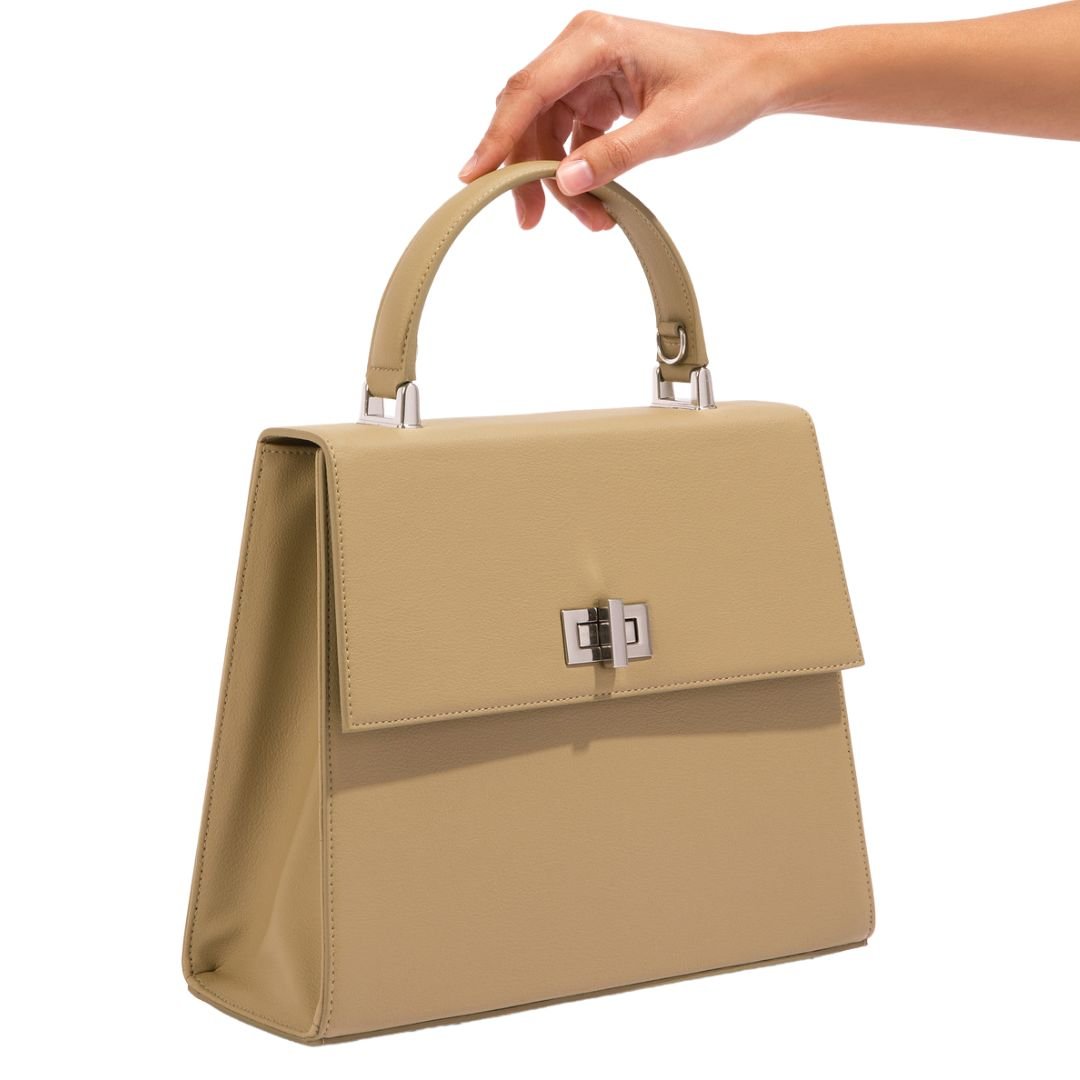 A person holding the beige She-EO handbag by its top handle, showcasing its compact yet elegant design