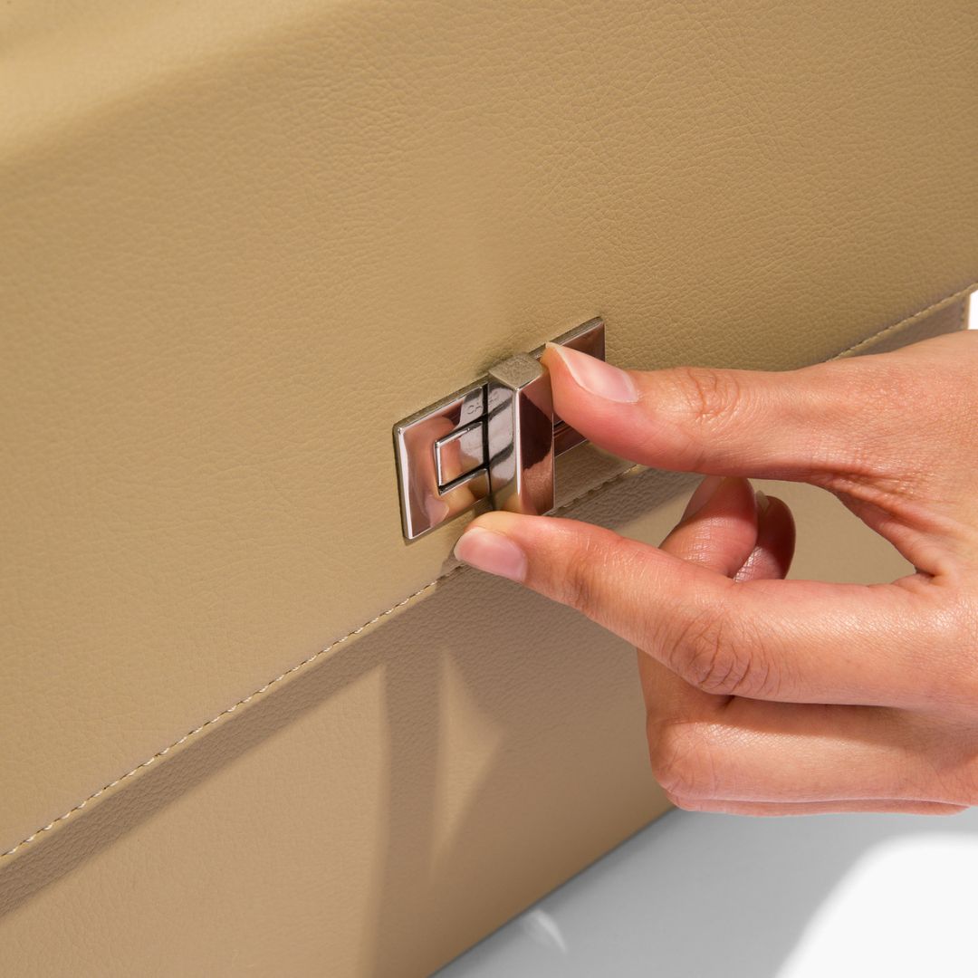 A close-up of a hand turning the silver lock on the beige She-EO handbag