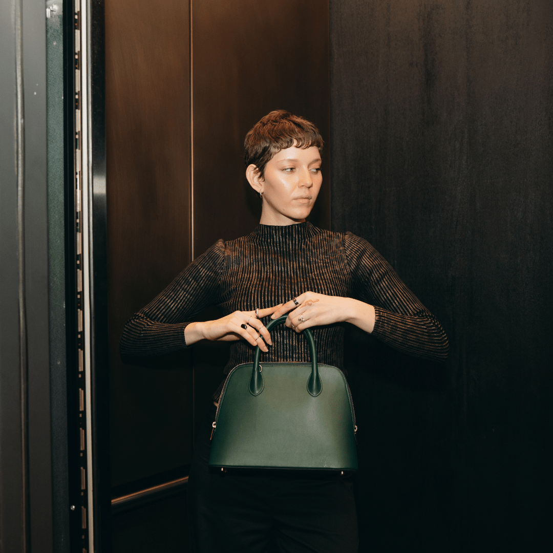 A stylish woman with short hair, dressed in a sleek black outfit, holds the Ms. Lady Luck handbag in an elevator with dark walls. She adjusts the bag’s handle while exuding confidence and sophistication.