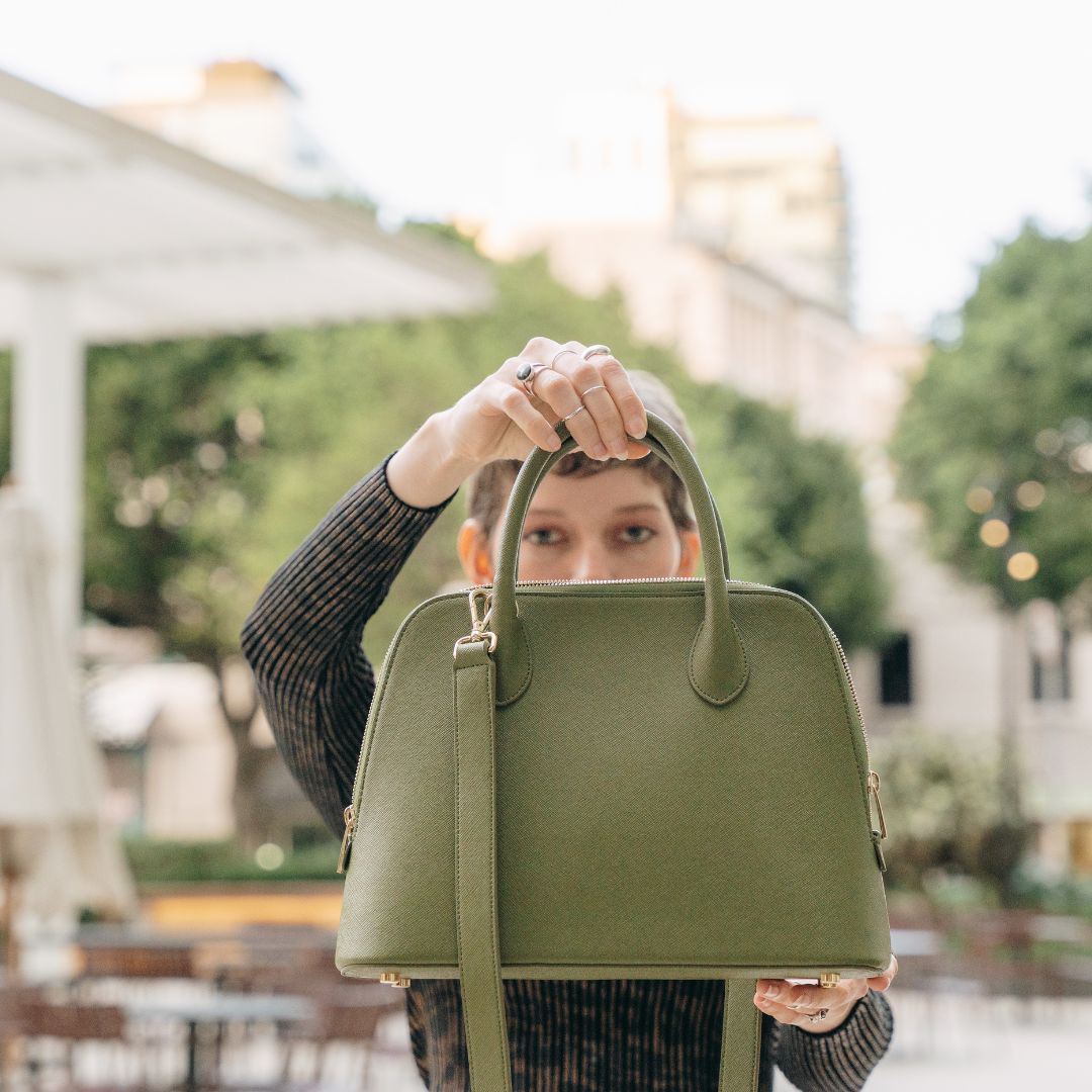 A woman holding the Ms. Lady Luck handbag in Olive Green, made from sustainable cactus leather. The structured silhouette and gold hardware shine against an urban backdrop, highlighting its elegant and eco-friendly design.