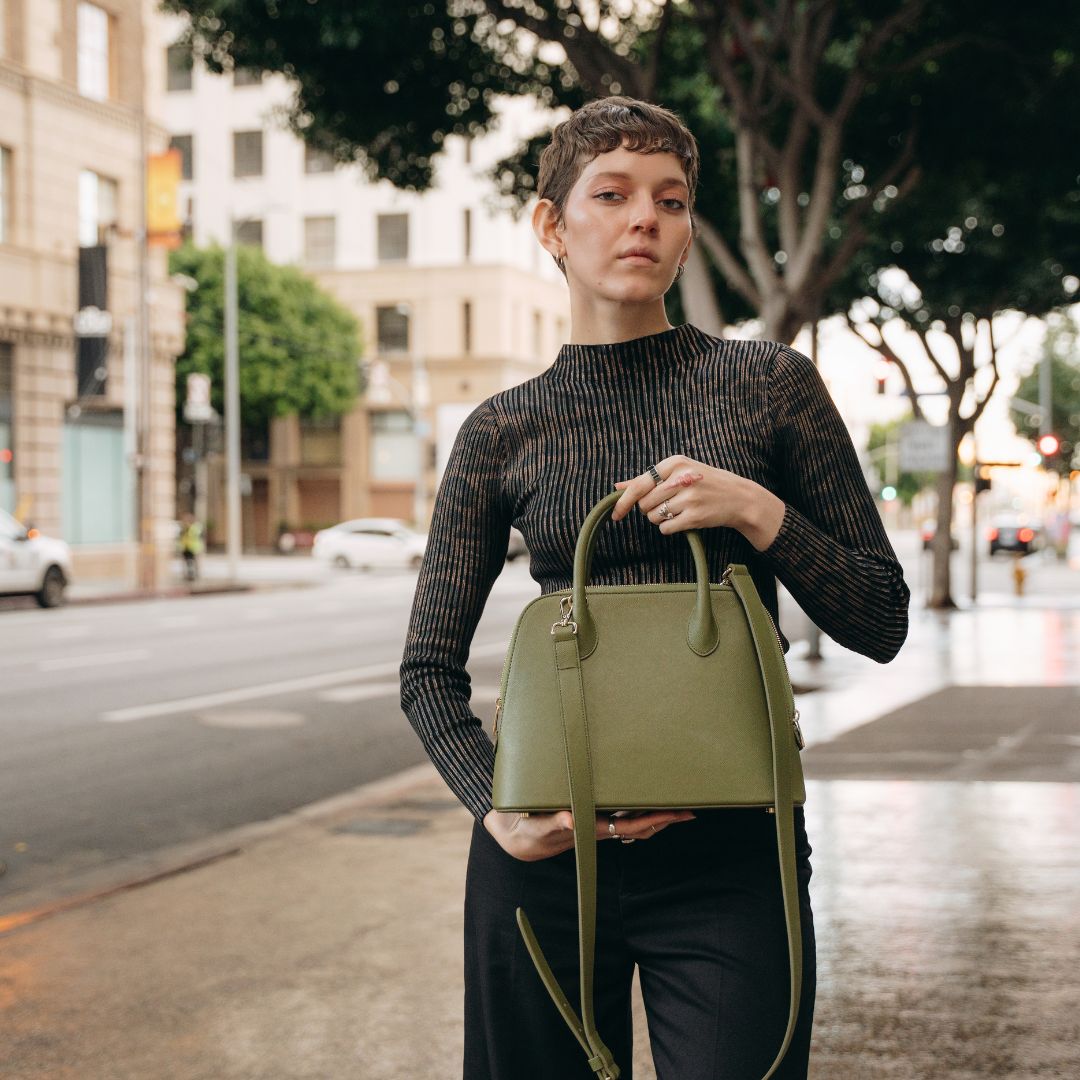A stylish woman holds the Ms. Lady Luck handbag in olive green, made from sustainable cactus leather, in an outdoor urban setting.