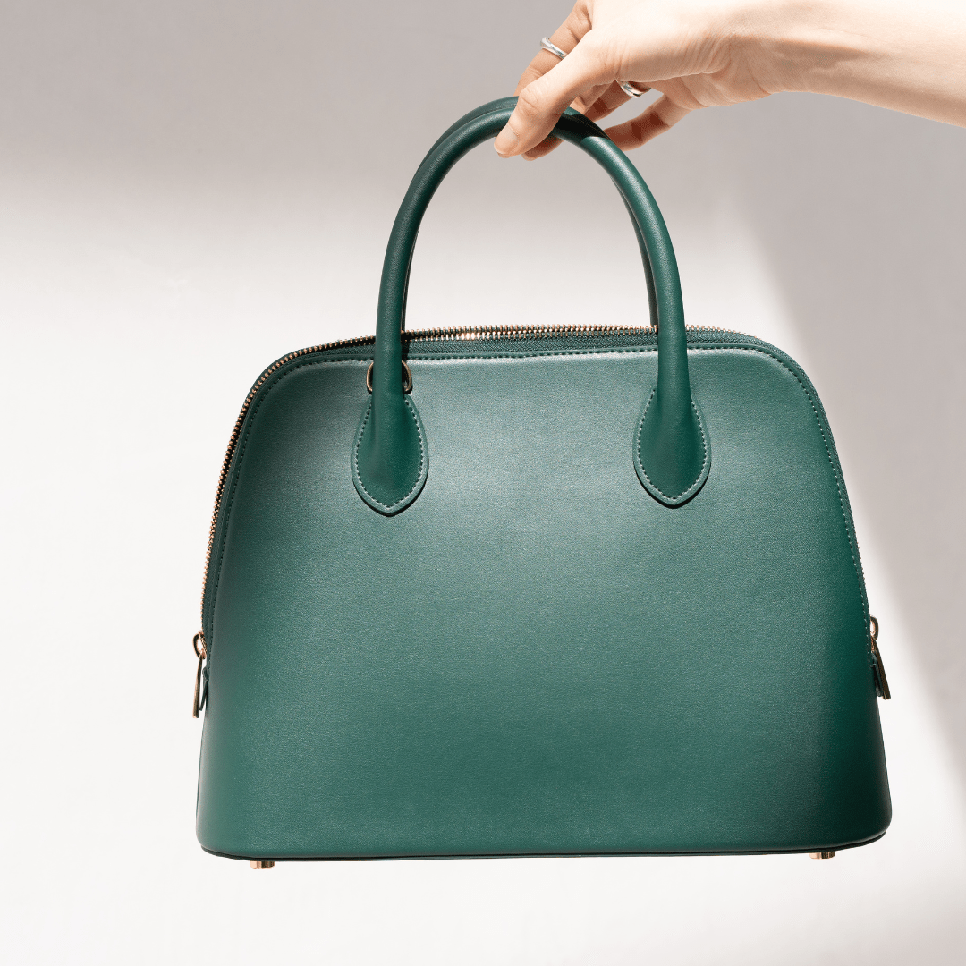 A close-up of a hand holding the Ms. Lady Luck handbag against a white backdrop. The rich green hue, fine stitching, and structured silhouette highlight the bag’s luxury craftsmanship.